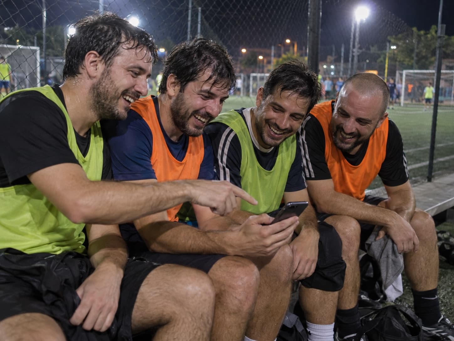 Cuatro amigos con pecheras, sentados al costado de la cancha, se ríen mirando una jugada en el celular